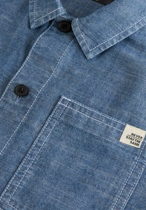 Camicia in chambray blu con colletto button-down, bottoni neri, taschino sul petto e un'etichetta con il testo "NON RIMANERE MAI UGUALE."