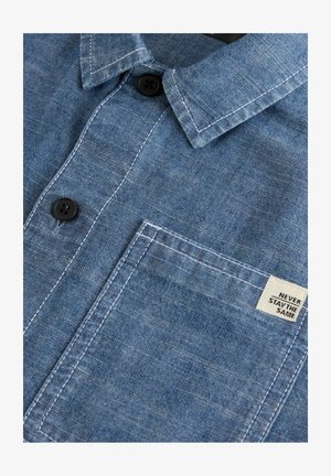 Cămașă din chambray albastru, cu guler cu butoni, butoane negre, un singur buzunar pe piept și o etichetă cu textul "NU RĂMÂNE NICIODATĂ ACELAȘI."