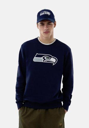 Junger Mann, der einen marineblauen Pullover und eine Mütze trägt, beide mit einem Seattle Seahawks-Logo, steht mit den Händen in den Taschen seiner olivgrünen Hose.