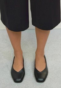 Mustad musta nahast plätud, millel on terav V-kujuline varvas, sile tekstuur ja minimalism, kantud mustade lühikeste pükstega.