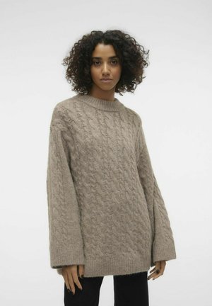 Kabelstrikket sweater i lys beige med rund hals, oversized ærmer og en blød tekstur. Sammen med sorte bukser.