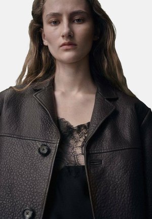 Giacca in pelle marrone con trama, con due bottoni neri, sovrapposta a un top in pizzo nero. Il design è oversized e contemporaneo.