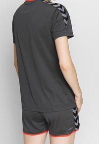 Conjunto de camiseta y pantalones cortos atléticos en gris con acentos rojos. Presenta un patrón de chevrón negro en los hombros. Fabricado con material transpirable.
