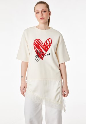 Giovane donna con pelle chiara e capelli raccolti che indossa una maglietta oversize color crema con un cuore rosso e la scritta "Love Endless". Gonna bianca con dettagli in pizzo.