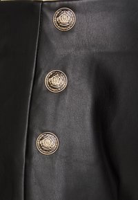 Vêtement en cuir noir avec trois boutons dorés ornés d'un design complexe de couronne et de laurier. Texture lisse avec une finition élégante.