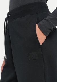 Melnas sporta bikses, izgatavotas no mīkstas auduma, ar elastīgu jostu un auklu, sānu kabatām un diskrētu logo uz augšstilba.