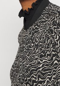 Bomber jacket s černobílým vzorem a texturovanou tkaninou, zdobený vlnitými liniemi a s celozipovým uzávěrem. Pod ním černý žebrovaný límec.