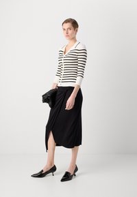 Cardigan rayé noir et blanc avec décolleté en V et fermeture à boutons, associé à une jupe portefeuille noire hauteur genoux et des escarpins noirs pointus.