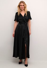 Rochie neagră, cu mâneci bufante scurte, talie legată și fenduire laterală. Material neted, asemănător cu satinul, până la gleznă. Pur tată cu tocuri înalte negre.