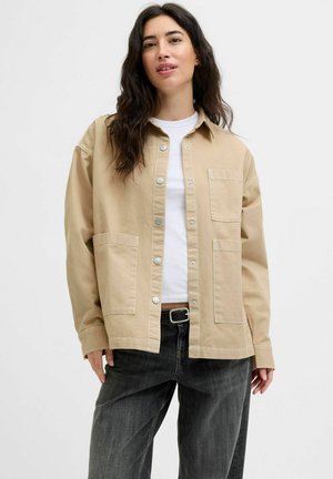 Vrouw met donker golvend haar, gekleed in een beige jasje met knopen over een witte blouse en losse zwarte jeans met een riem, staande tegen een effen achtergrond.