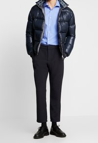 Marinblå pufferjacka med quiltad textur, grå dragkedja och mönstrad foder; ljusblå skjorta; mörka skräddarsydda byxor; svarta skor.