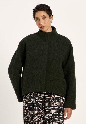 Woman with short curly hair wearing a dark green textured high-neck sweater and black and beige patterned pants standing against a light background.