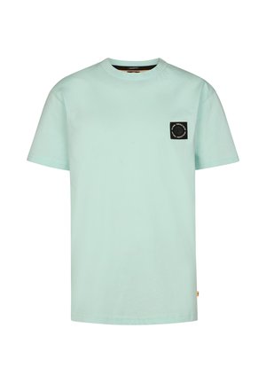 Jongen staat recht, kijkt naar voren, draagt een lichtblauw T-shirt met een klein zwart logo, zwarte broek en witte sneakers tegen een effen achtergrond.