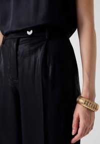 Schwarze, hoch taillierte Hose mit glänzendem Finish, gesichert durch einen silbernen Knopf, kombiniert mit einem goldfarbenen, gerippten Armband an einem nackten Arm.