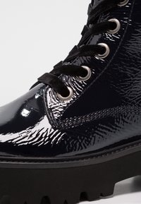 Bottine en cuir verni noir avec une finition texturée, œillets en argent, lacets noirs et une semelle en caoutchouc chunky avec un profil profond pour une meilleure adhérence.
