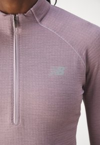 Nærbillede af person iført en mauve kvart-zip sportsbluse med New Balance-logo på brystet, der viser detaljer ved halsudskæring og skulder.