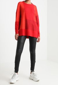 Pull oversize rayé rouge et orange avec une finition texturée, associé à des leggings noirs ajustés avec des détails côtelés, et des baskets blanches.