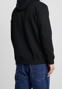 Sweat à capuche noir avec cordon de serrage et poignets côtelés, associé à un jean en denim bleu avec des détails de couture sur les poches arrière.