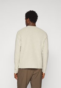 Pull en maille texturée, beige clair, avec des manches longues, un col rond et une coupe décontractée, associé à un pantalon marron foncé. Détails du motif tricoté visibles.