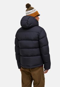 Mand iført en marineblå dunjakke med hætte, brune bukser og en brun-hvid strikket hue med en pom-pom, set fra bagsiden mod en ensfarvet baggrund.