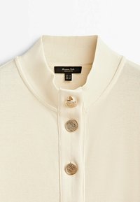 Polo lavorato a maglia crema con colletto alto, caratterizzato da tre bottoni decorativi dorati e un'etichetta interna che indica la taglia e l'origine.