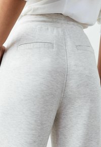 Hellgraue Sweatpants aus weichem, strukturiertem Stoff mit zwei Gesäßtaschen, die ein dezentes Streifenmuster und abgestepptes Detail aufweisen.
