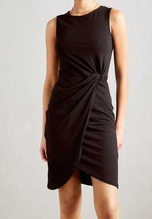 Ärmelloses schwarzes Kleid mit rundem Halsausschnitt und drapiertem asymmetrischem Wickeldetail vorne, getragen von einer stehenden Person vor neutralem Hintergrund.