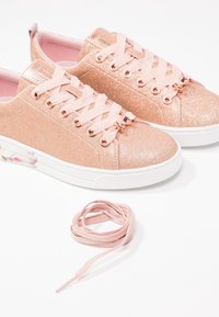 Zapatillas brillantes en oro rosa con cordones de satén rosa, suela de goma blanca y detalle floral transparente en el talón. Se muestra un cordón adicional.