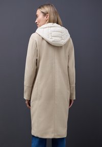Beige Mantel mit gepolsterter weißer Kapuze, maßgeschneiderter Schnitt, langen Ärmeln und geradem Schnitt. Glatte Textur, minimale Details auf dem Stoff.