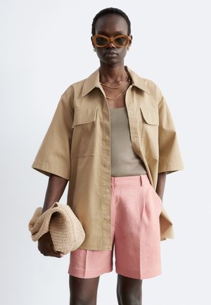 Dunkelhäutige Person trägt übergroße beige Jacke, taupefarbenes Tanktop, pinke Shorts, große braune Sonnenbrille, goldene Halsketten, hält eine beige gestrickte Clutch.