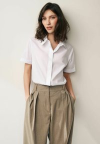 Chemise blanche à manches courtes avec un col, associée à un pantalon beige à rayures verticales taille haute, doté de plis à l'avant et de poches latérales.