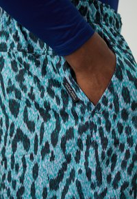 Teal ir juodų leopardų rašto sportiniai šortai su elastingu juosmeniu ir šoninėmis kišenėmis. Matoma prekės etiketė kišenėje.