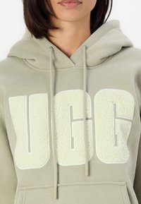 Gaiši zaļa dvidaļa ar mīkstu audumu, kurai ir liels, pūkains "UGG" logos krēmkrāsā. Iekļaujās izvelkamās auklas un priekšējais kabata. Ikdienišķs dizains.