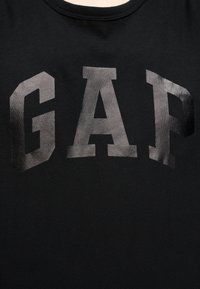 Μαύρο ύφασμα με μεγάλα, έντονα γκρίζα γράμματα που σχηματίζουν τη λέξη "GAP" στην περιοχή του στήθους.