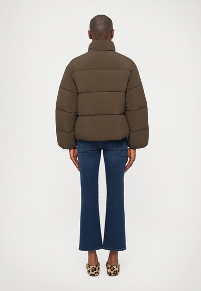 Giubbotto imbottito in verde oliva scuro con colletto alto, design trapuntato e silhouette voluminosa, abbinato a jeans blu a zampa d'elefante e scarpe con stampa animalier.