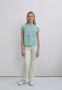 T-shirt bleu clair court avec un graphique coloré de tête de mort et os croisés, associé à un pantalon évasé blanc cassé et des baskets blanches.