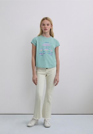 Light blue cropped t-shirt featuring a colorful skull and crossbones graphic, paired with off-white flared pants and white sneakers.