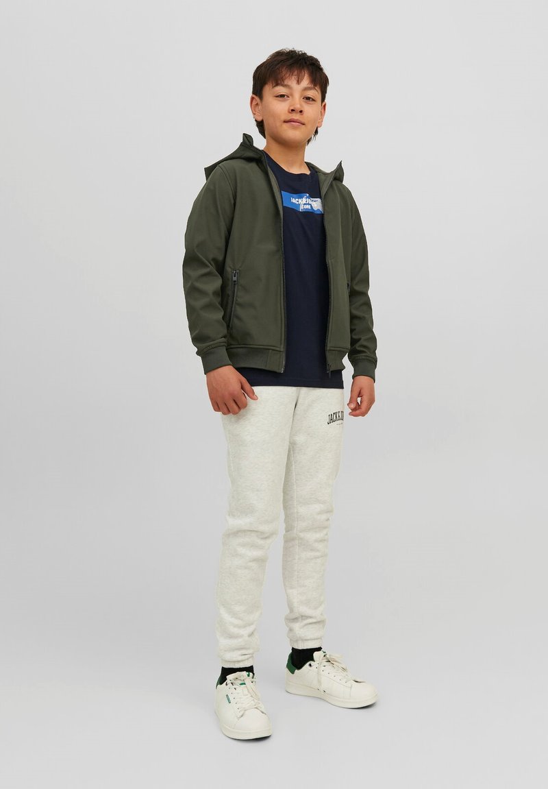 Grøn zip-up hoodie, navy t-shirt med logo, lysegrå sweatpants og hvide sneakers med grønne accenter. Model står foran en ensfarvet baggrund.