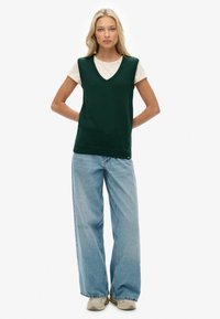 Groene gebreide vest met een V-hals, gedragen over een witte t-shirt, gecombineerd met lichtblauwe wijdvallende jeans en tan sneakers.