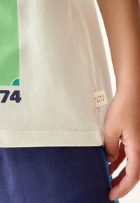 Baltas medvilninis marškinėlis su žalia grafine spauda, vaizduojančia numerį 74. Šoninė etiketė su užrašu „Little Bird“. Dėvimas su tamsiai mėlynomis kelnėmis.