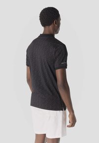Homme de dos portant un polo à manches courtes noir moucheté et un short blanc, sur un fond clair uni.