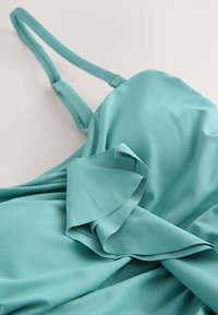 Robe sans manches bleu sarcelle clair avec bretelles réglables et détail de volants superposés sur le corsage, posée à plat sur une surface blanche.