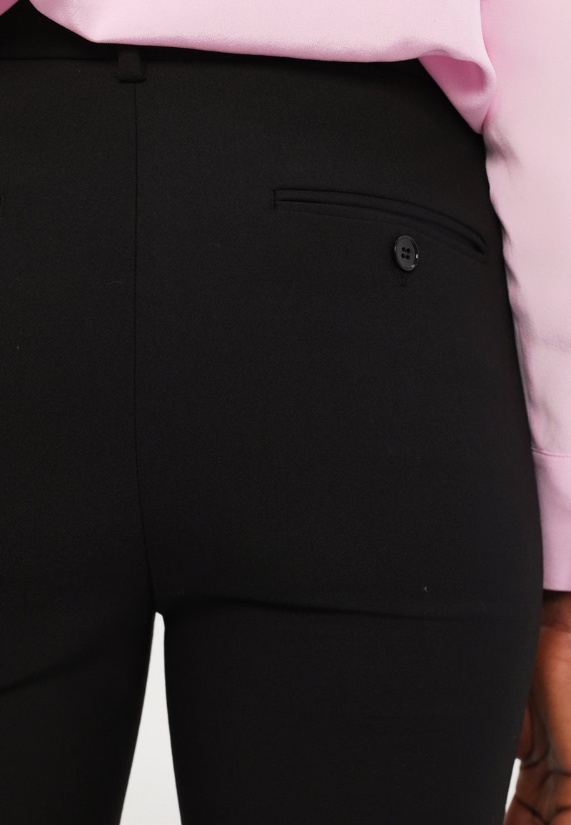 Czarne spodnie o kroju tailorskim z tylną kieszenią zapinaną na guzik oraz gładką, matową fakturą, w połączeniu z jasnoróżową bluzką częściowo widoczną.