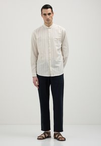 Camicia lunga beige chiaro a maniche lunghe con righe verticali e colletto a banda, abbinata a pantaloni larghi blu scuro e sandali marroni con cinturini.