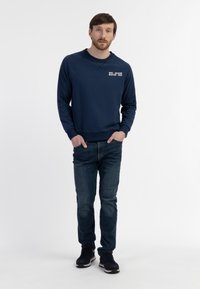 Marinblå sweatshirt med rund hals og ribbede manchetter, med hvid tekst på venstre bryst. Sammen med mørke jeans og sorte sneakers.