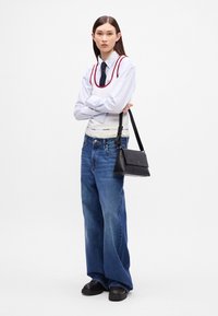 Modelka nosi białą koszulę w paski, tank top z czerwoną lamówką, oversize'owe niebieskie dżinsy i czarne buty, trzymając małą czarną torbę na ramię.