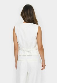 Vit ärmlös väst med ett bälte, som har en mjuk textur och rena linjer, sedd bakifrån.