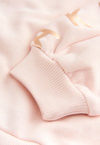 Manche de sweat-shirt rose doux avec poignet côtelé et texte partiellement imprimé en doré brillant sur le tissu.