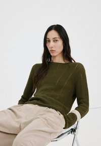 Kvinde med langt mørkt hår siddende på metalstol, iført olivengrøn strikket trøje og beige bukser med vide ben mod ensfarvet hvid baggrund.