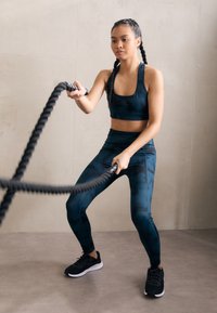Cuerdas de entrenamiento negras sostenidas por una persona que lleva un sujetador deportivo y leggings de diseño teal, de pie sobre un suelo gris texturizado.